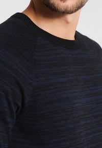 Granatowy sweter dzianinowy z teksturowanym wzorem, czarnym ściągaczem przy szyi oraz rękawami raglanowymi, ukazujący połączenie ciemnych i jaśniejszych odcieni.