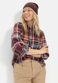 Rutete flannel skjorte i varme nuancer af rød, brun og grå med knapper og opfoldede ærmer, parret med khaki bukser og en mørkerød hue.