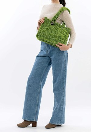 Femme en pull beige et jean à jambes larges, tenant un grand sac à main vert texturé sur l'épaule, portant des bottines marron à talons.