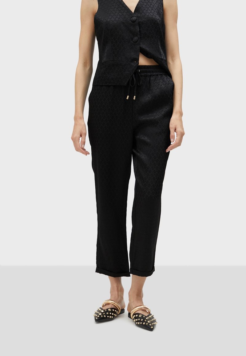 Femme portant un gilet noir à boutons, un pantalon noir assorti à cordon de serrage, et des mules noires à bout pointu avec des picots dorés et des brides aux chevilles, se tenant sur un sol gris.