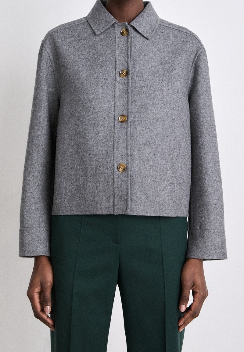Personne portant une veste en laine grise boutonnée avec un col et un pantalon vert foncé sur mesure, sur un fond uni.