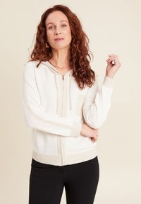 Cardigan à capuche blanc en tissu tricot texturé, doté d'un zip sur le devant, d'une petite poche poitrine et de détails beiges contrastants au niveau des poignets et de l'ourlet.