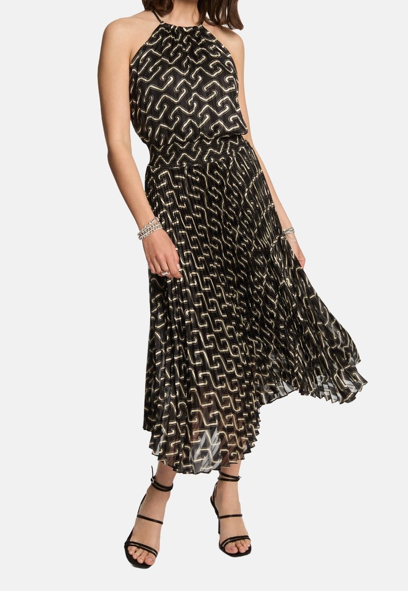 Femme portant une robe noire sans manches à col halter avec un motif géométrique blanc, jupe plissée, bracelets argentés et talons noirs à lanières.