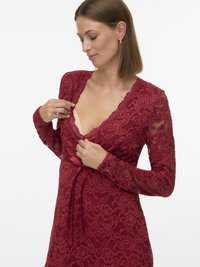 Γυναίκα που φοράει κόκκινο δαντελένιο φόρεμα με μακριά μανίκια και λουλουδάτα σχέδια, διορθώνοντας το βαθύ V-λαιμόκοψη για να αποκαλύψει ένα μπεζ σουτιέν από κάτω.