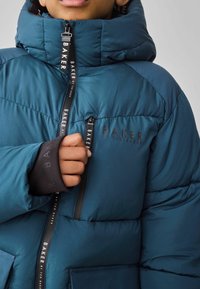 Doudoune teal avec un col montant, dotée de zippers noirs, de poches et d'accents de logo. Le matériau a une finition lisse et texturée.
