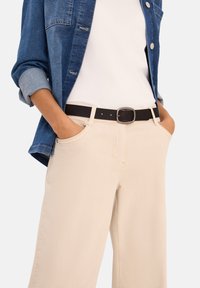 Hellbeige weite Hosen mit Fronttaschen, kombiniert mit einem weißen T-Shirt und einem blauen Jeanshemd. Ein schwarzer Gürtel mit einer quadratischen Schnalle rundet den Look ab.