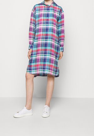 Sukienka koszulowa z bawełnianej tkaniny w kratę, łącząca kolory różowy, niebieski i zielony, z długimi rękawami, kołnierzykiem i rozcięciem po bokach. Noszona z białymi trampkami.