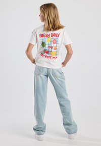 Camiseta blanca de algodón con un gráfico colorido de frutas y texto rojo en la parte posterior. Combinada con jeans holgados de color azul claro y zapatillas blancas.