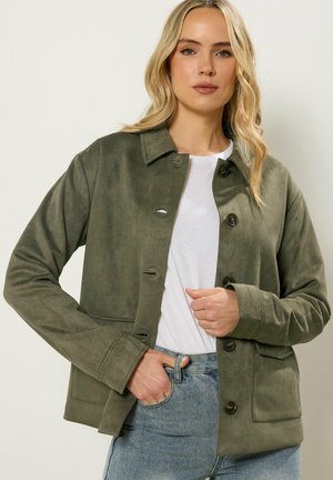 Blonde vrouw die een olijfgroene jas met knopen draagt over een wit shirt en lichtblauwe jeans, poserend met één hand in de zak.