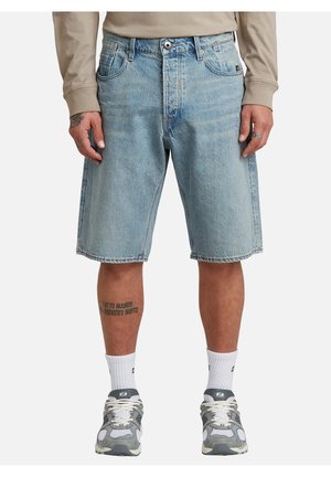 Mann trägt hellblaue Jeansshorts, beiges Langarmshirt, weiße Socken und graue Turnschuhe, steht mit entspannten Händen an den Seiten.