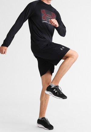 Persona vestida con ropa deportiva negra de Under Armour y zapatillas, levantando una rodilla con los brazos doblados, sobre un fondo blanco liso.