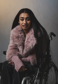 Cappotto di pelliccia finta rosa con ampio collare, maniche rosa a coste e pantaloni neri luccicanti. Modella su una sedia a rotelle con lunghi capelli scuri.