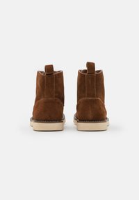 Botas de tobillo de ante marrón con una textura suave, costuras blancas en contraste y una lengüeta en la parte posterior. Suela de goma de color crema.