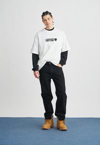Weißes Oversize-T-Shirt mit grafischem Druck, darüber getragen ein schwarzes Langarmshirt. Schwarze Hose und beige Arbeitsstiefel komplettieren das Outfit.