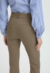 Pantalones marrones ajustados con una textura suave, cintura a media altura y dos bolsillos traseros. Combinados con una blusa rayada en azul y blanco.
