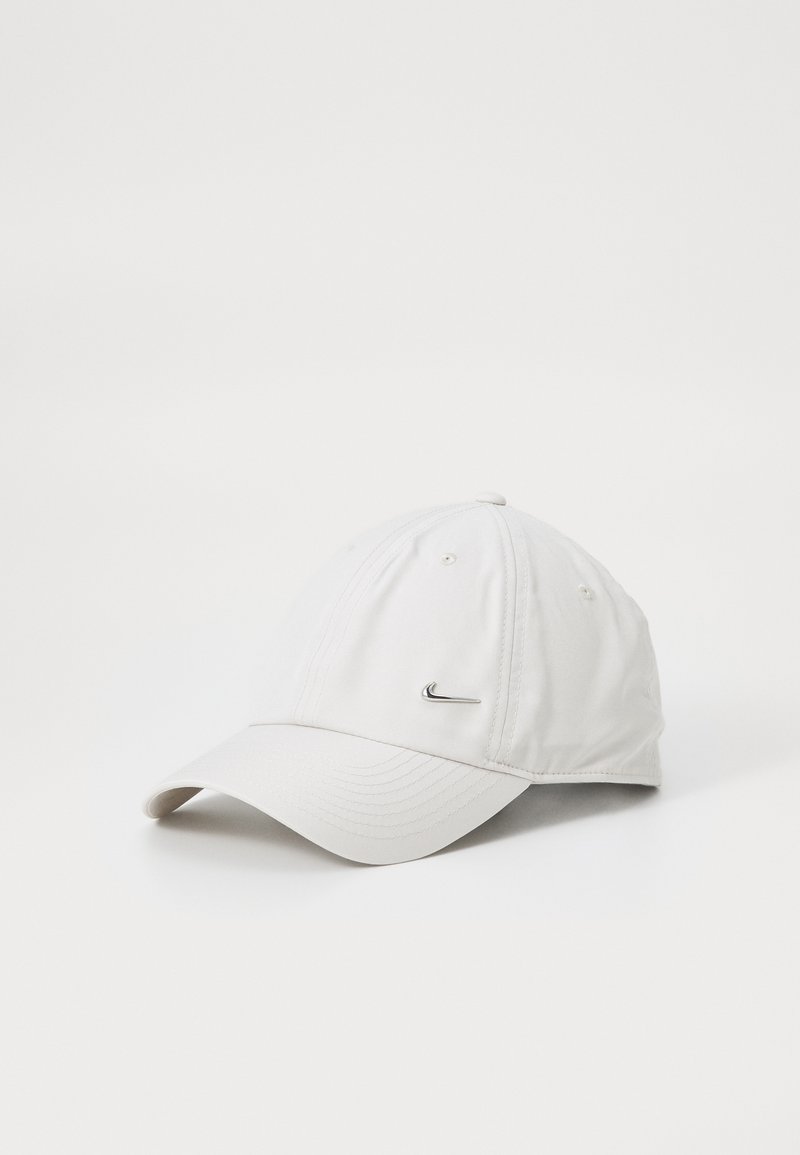 Biely baseballový klobúk s malým kovovým logom Nike swoosh vpredu, vystavený na jednoduchom bielom pozadí.