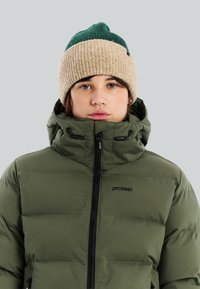 Jongere die een groen en beige gebreide muts en een gesloten olijfgroene gewatteerde jas met capuchon draagt, tegen een effen achtergrond.