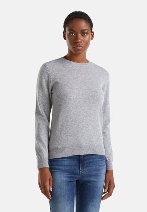 Woman wearing a light gray long-sleeve knit sweater and blue jeans, standing against a plain white background, facing forward.