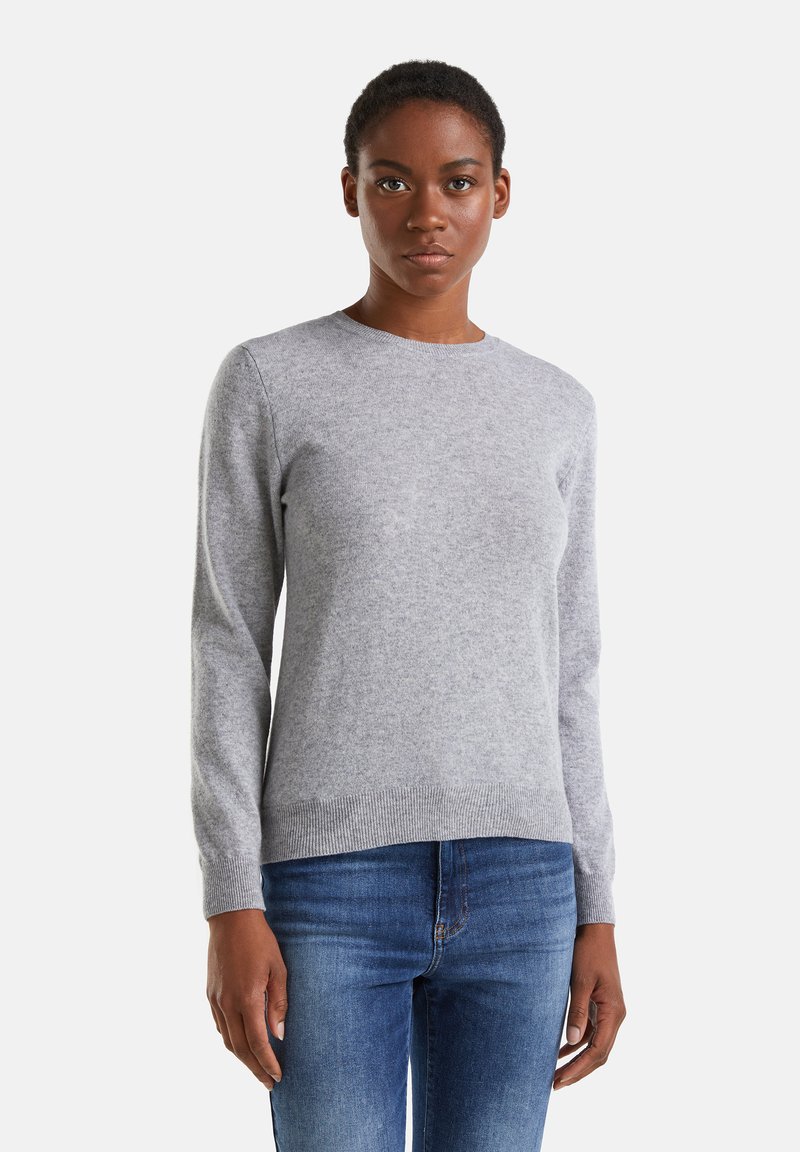 Kobieta w jasnoszarym swetrze z długim rękawem i niebieskich dżinsach, stojąca na tle gładkiej białej ściany, zwrócona w stronę obiektywu.