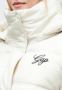 Hvit boblejakke med høy krage, trykknapper og svart brodert "Gap"-logo på brystet. Myk tekstur og quiltet design.