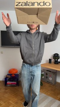 Cardboard box with "zalando" logo held above a person in a gray hoodie and light blue jeans, standing in a room with diverse furniture.