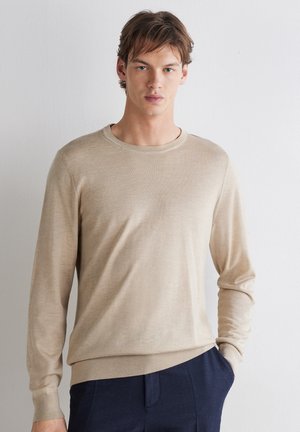 Ung mand står op ad en hvid væg iført en beige langærmet sweater og mørkeblå bukser med den ene hånd i lommen.