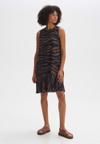 Ärmelloses Kleid aus schwarzem Stoff mit einem braunen Tigerstreifenmuster, das einen entspannten Schnitt und einen Rüschensaum aufweist. Kombiniert mit braunen Sandalen.
