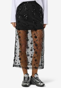 Gonna nera in pizzo con motivo floreale in paillettes, sovrapposta a una fodera interna più corta, abbinata a sneaker sportive grigie e nere.