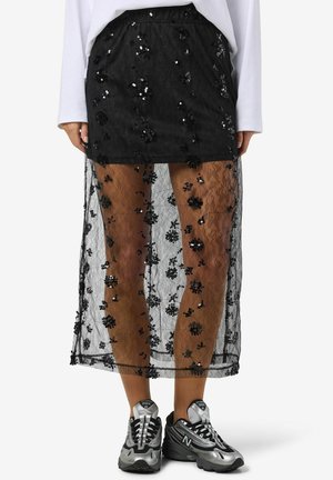 Zwarte kanten rok met een bloemig paillettenpatroon, gelaagd over een kortere binnenvoering, gecombineerd met grijze en zwarte sport sneakers.
