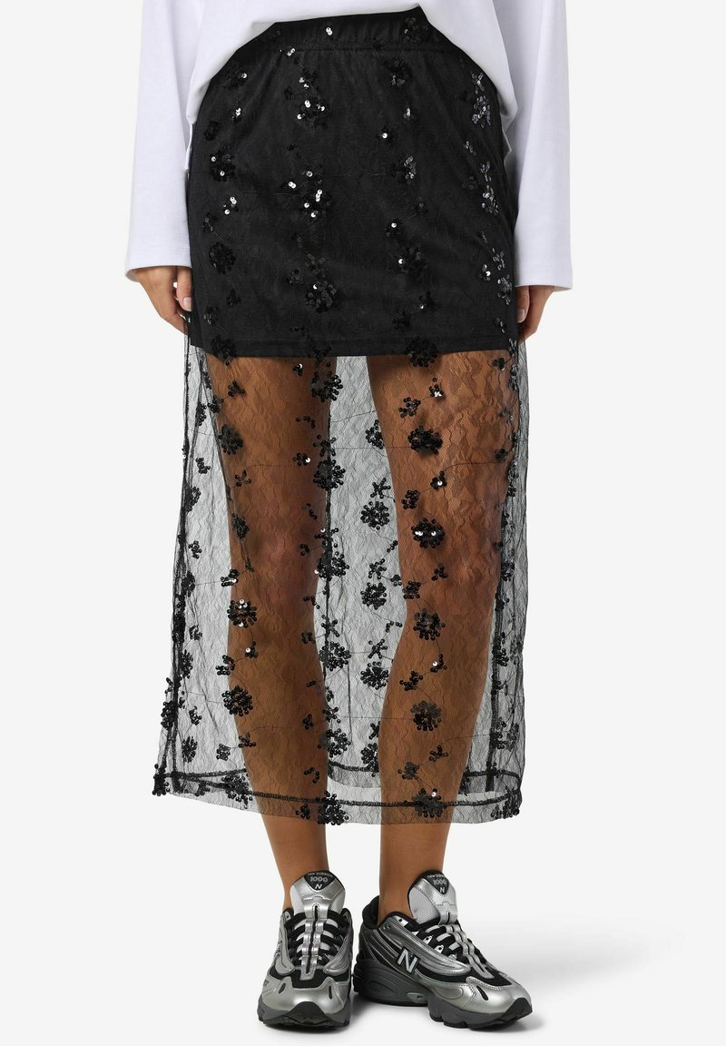 Gonna nera in pizzo con motivo floreale in paillettes, sovrapposta a una fodera interna più corta, abbinata a sneaker sportive grigie e nere.