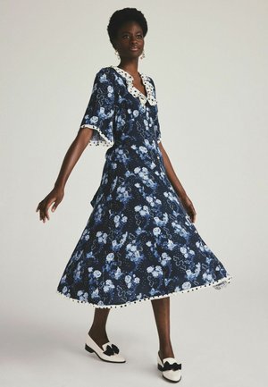 Marine bloemenjurk met blauw en witte patronen, gerimpelde stippenkraag, korte mouwen en pomponrand aan de zoom; gecombineerd met zwart-witte schoenen.