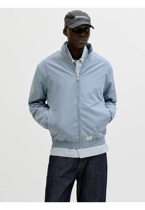 Mann trägt hellblaue Zip-Up-Jacke, kariertes Hemd, dunkle Jeans und graue Kappe mit "JACK & JONES"-Logo, steht mit den Händen in den Taschen.