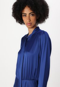 Blusa blu in satin con scollatura alta, spalle plissettate e vita elasticizzata. Tessuto liscio e maniche lunghe.