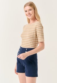 Lichtbruin geribbeld topje met witte strepen, korte mouwen. Gecombineerd met een hoogdoorgetrokken marineblauwe broek met twee zilveren knopen aan de voorkant.