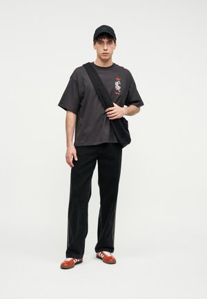 Νέος άνδρας που φοράει μαύρο καπέλο, φαρδιά μαύρη μπλούζα με γραφικό σχέδιο, μαύρο παντελόνι, κόκκινα και λευκά αθλητικά παπούτσια και μαύρη τσάντα χιαστί, στέκεται μπροστά σε λευκό φόντο.