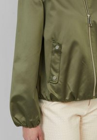 Veste bomber vert olive avec une fermeture éclair, des poignets élastiques et une poche latérale avec fermeture à pression. Texture lisse avec une finition légèrement brillante.