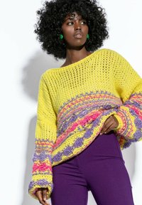 Gruby żółty sweter dziany z fioletowymi i różowymi akcentami, z szerokimi rękawami i luźnym krojem; zestawiony z obcisłymi fioletowymi spodniami.