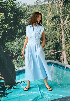 Žena v svetlomodrých šatách bez rukávov a čiernych žabkách stojaca pri bazéne s hustými zelenými stromami v pozadí, usmievajúca sa a pozerajúca sa do strán.