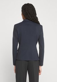 Blazer bleu marine à coupe ajustée, épaules structurées et fente unique au dos. Tissu lisse et détails de couture minimalistes. Associé à un pantalon foncé.