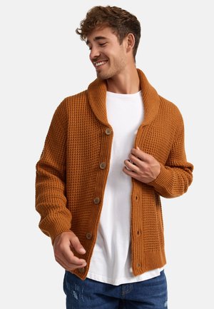 Rustfarget strikket cardigan med sjalkrage, fem knapper foran og ribbestrikkede mansjetter. Brukt over en hvit t-skjorte.
