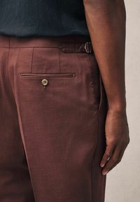 Brown tailored trousers with a smooth texture, featuring a buttoned back pocket and side adjusters. Hand resting on the hip area.