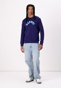 Navy sweatshirt with "GAP NYC" text in light blue, paired with light blue jeans and gray sneakers. Smooth texture, relaxed fit.