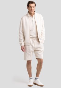 Lys beige zip-jakke over en matchende polo og shorts. Hvide sneakers med brune såler. Blå- og hvidstribede sokker.