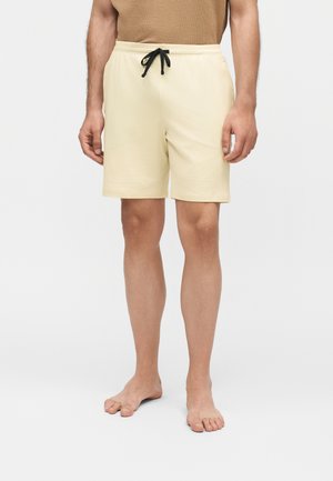 Homme portant un short beige à cordon et un haut marron, debout pieds nus devant un fond clair uni.