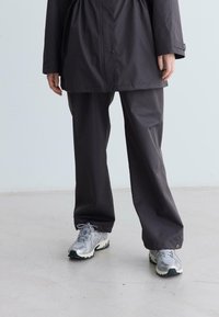 Eine Person trägt lockere, dunkle Hosen und silberne Sportschuhe und steht auf einem hellen Boden vor einer schlichten Wand.