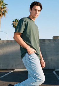 Noor mees tumedate juustega, kes kannab oliivirohelist T-särki ja helesiniseid teksapükse, seisab väljas halli tellismüüri lähedal, üks käsi püksitaskus.