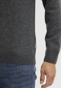 Pull gris en maille avec ourlet et poignets côtelés, fabriqué en tissu doux. Une petite étiquette est visible en bas à gauche. Porté avec un jean bleu.