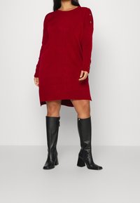 Robe en maille rouge avec encolure ronde et manches longues, ornée de boutons sur l'épaule et d'un ourlet asymétrique. Associée à des bottes noires montantes.