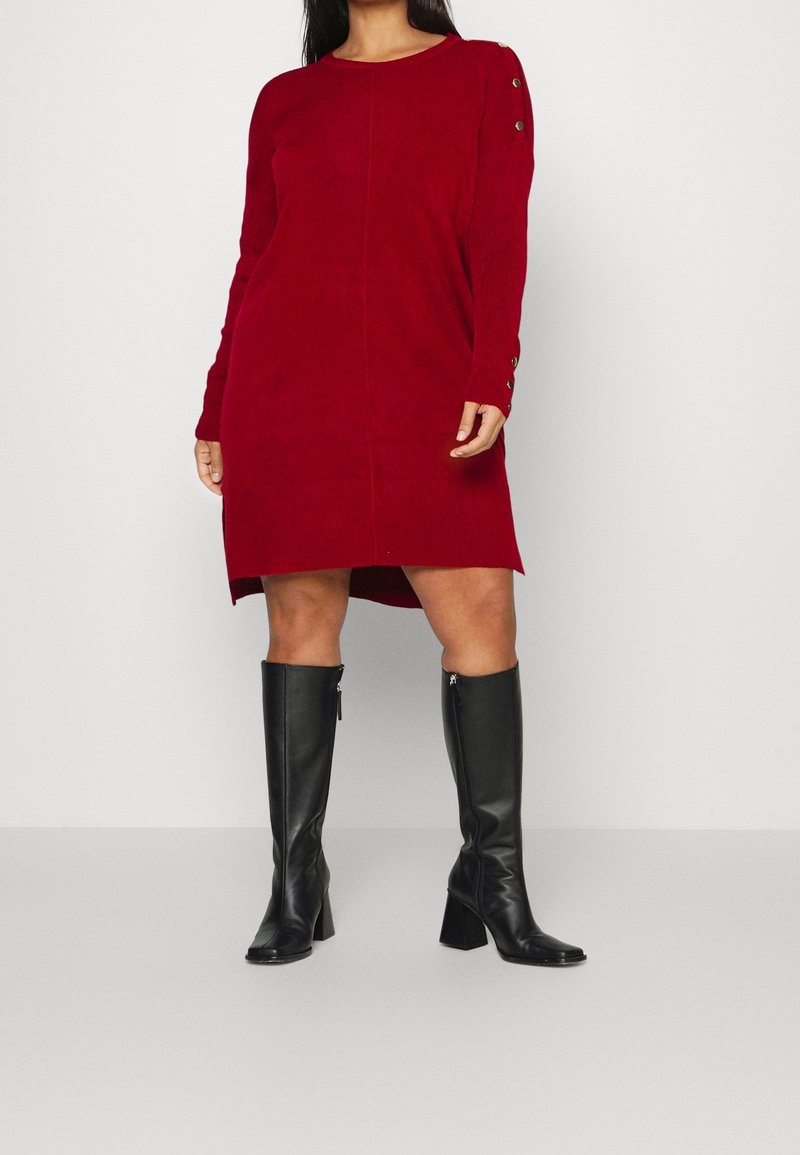 Robe en maille rouge avec encolure ronde et manches longues, ornée de boutons sur l'épaule et d'un ourlet asymétrique. Associée à des bottes noires montantes.