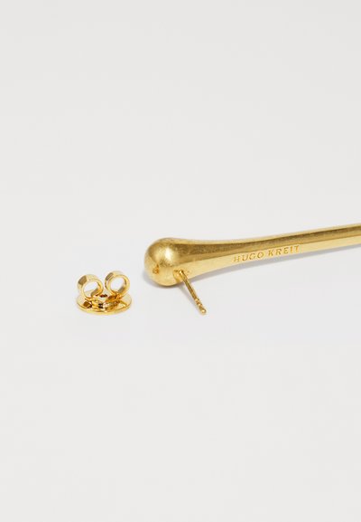 Conjunto de joyería de oro que presenta un pendiente largo y curvado con una textura suave y el nombre "HUGO KREIT" grabado, junto a un respaldo circular.